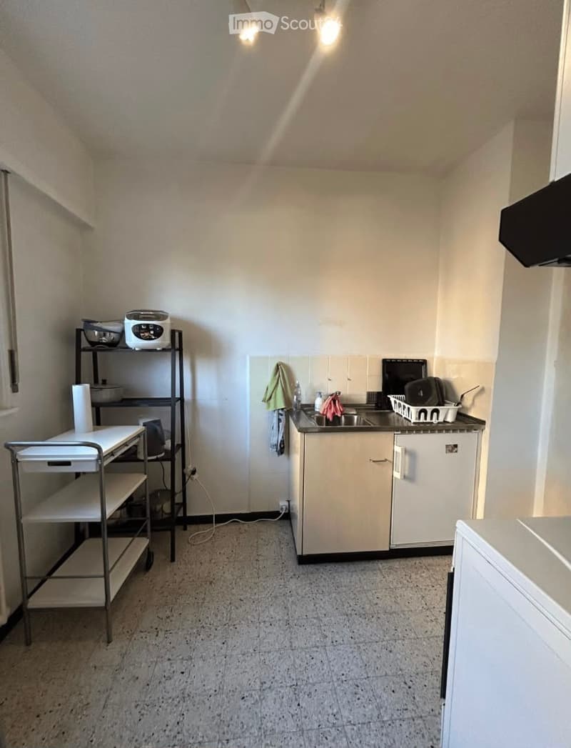A small kitchen with white cabinets, a sink, a microwave, an oven, a cart, and a refrigerator. The floor is tiled, and there are various kitchen items on the counter and shelves.