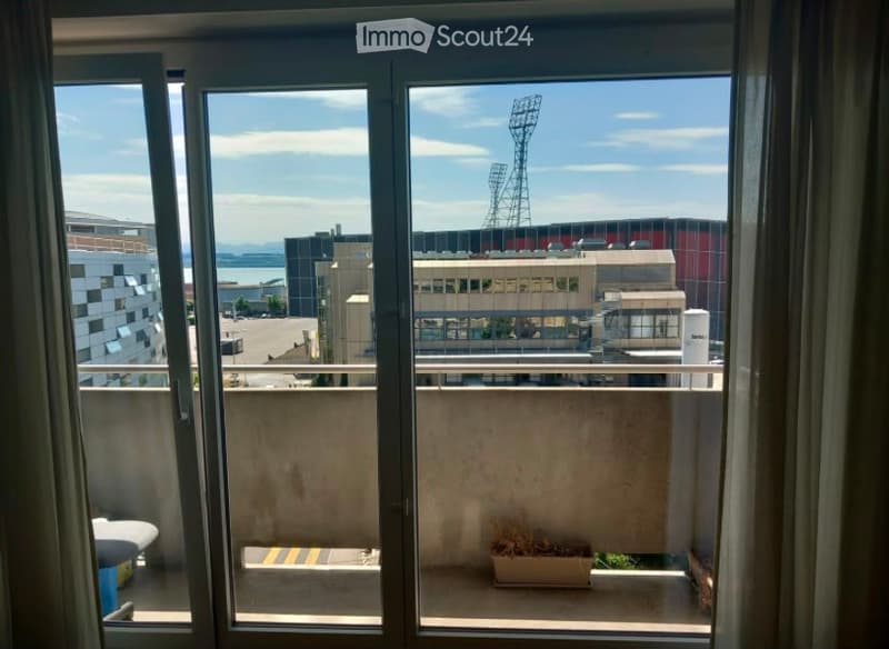 Balcony with a view of a cityscape, including a stadium and surrounding buildings. The balcony has a metal railing and a small potted plant.
