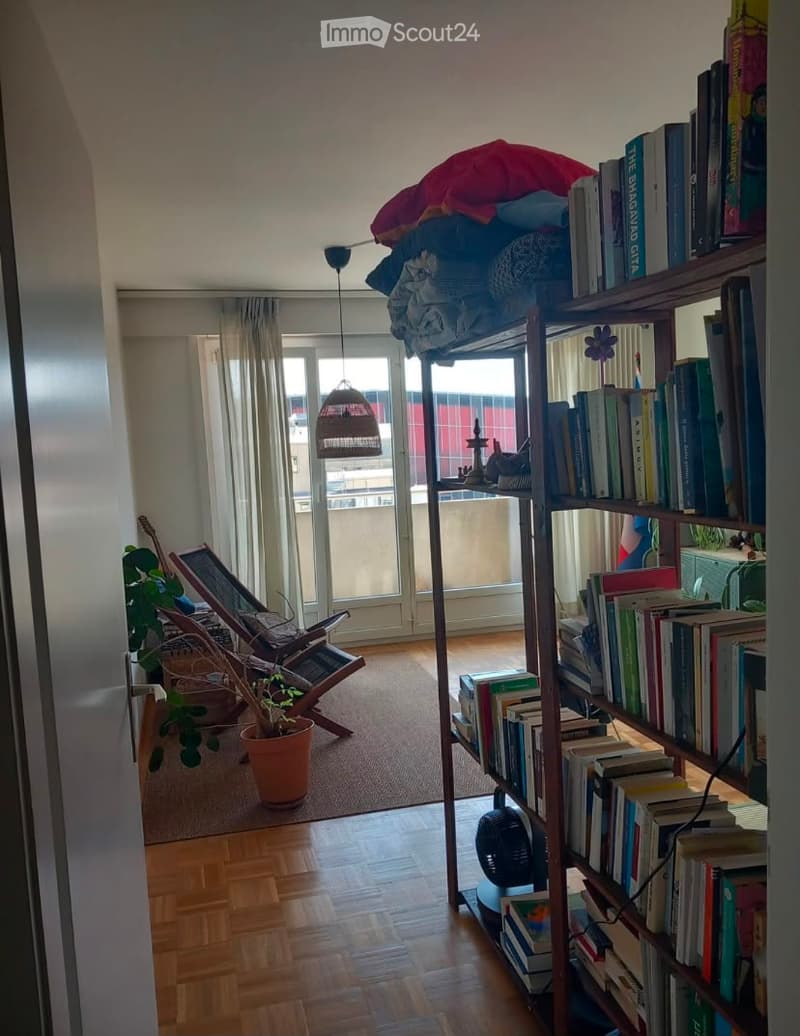 A cozy, well-lit room with wooden flooring, a large bookshelf filled with books, a wicker hanging basket, a potted plant, a wooden chair, and a guitar leaning against the wall. The room has large windows with sheer curtains and a view of a balcony.
