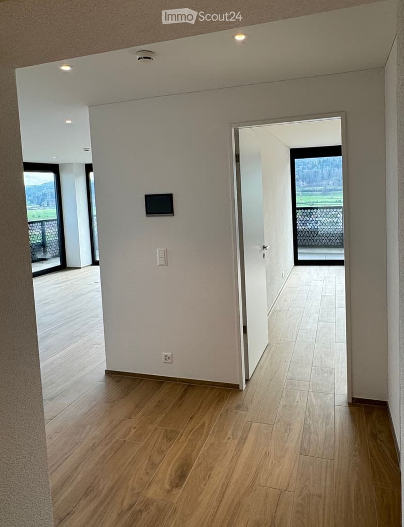 Modern hallway with light wooden flooring, white walls, recessed lighting, and a small screen mounted on the wall. The hallway leads to an open area with large glass doors offering a scenic view.
