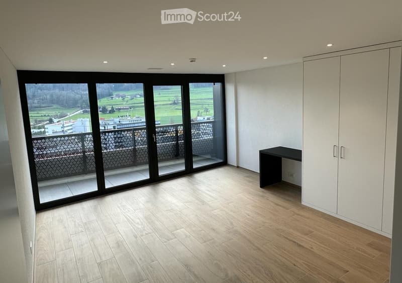 Empty room with wooden flooring, large sliding glass doors leading to a balcony, white built-in wardrobe, and a small black table.