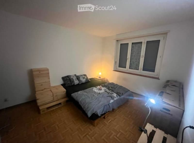 Cozy bedroom with wooden flooring, a double bed with grey bedding, a nightstand with a lamp, and a wooden crate on the left side.