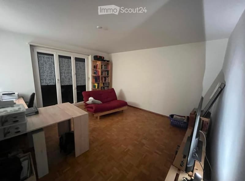 A small, well-lit room with wooden flooring, a red sofa, a wooden table with a chair, a bookshelf, and a large window with a view of the balcony.