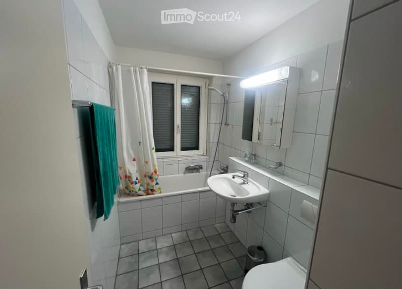 A small, tiled bathroom with a bathtub, sink, mirror, and toilet. A towel hangs on the wall, and a shower curtain is visible in the bathtub area.