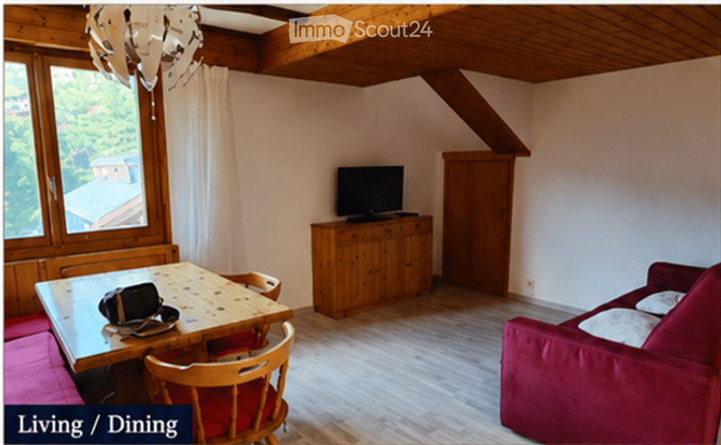 Living/dining area with wooden floors, large window with view, red sofa, wooden dining table with chairs, TV on wooden cabinet, and exposed wooden ceiling beams.