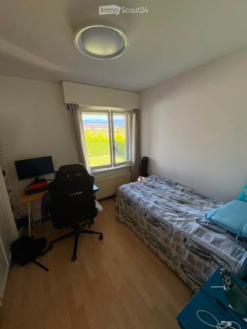 A small, well-lit bedroom with a single bed, a desk with a computer, and a gaming chair. The room features wooden flooring and large windows with a scenic view.