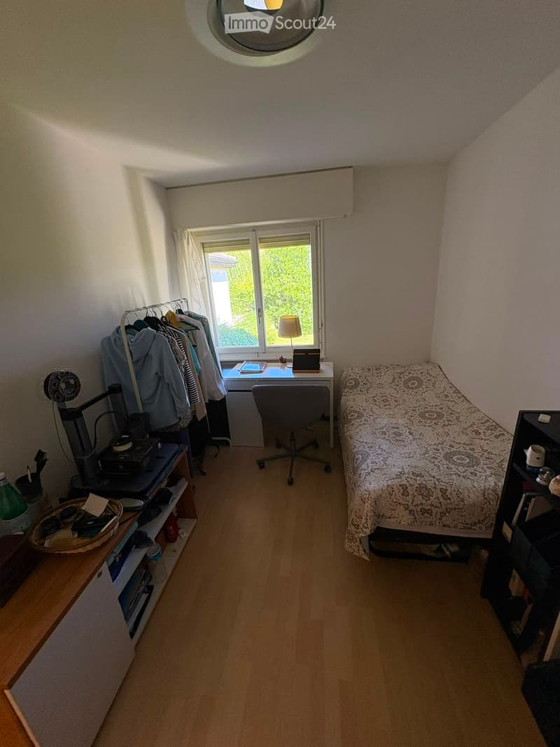 Cozy bedroom with wooden flooring, a bed with patterned bedspread, a desk with a chair, a wardrobe, and a shelf with various items.