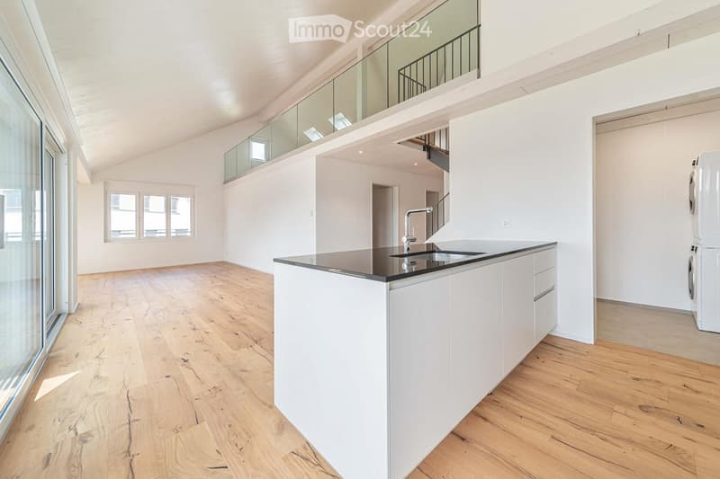 Open-plan space with light wooden flooring, white cabinetry, and a modern kitchen island. The room features large windows and a balcony area with glass railings.