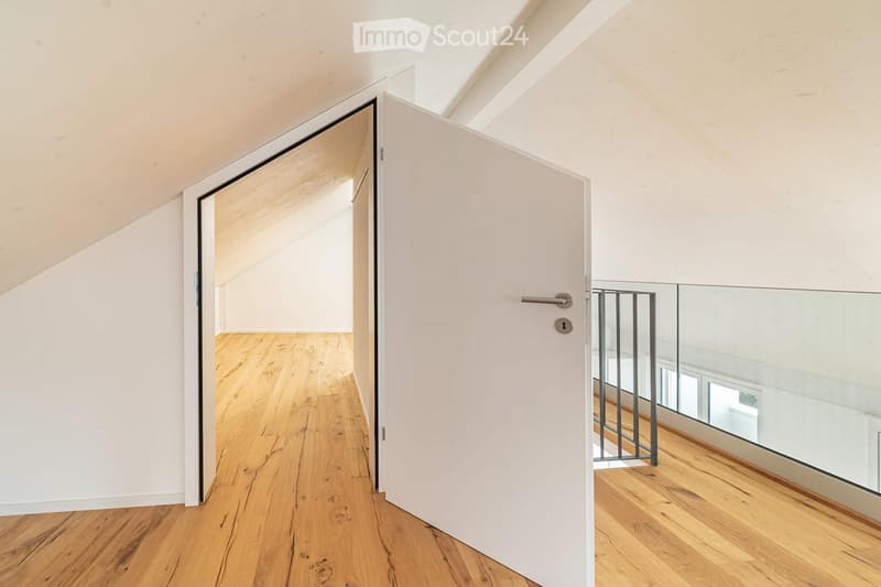 A modern, minimalist hallway with light wooden flooring, white walls, and a glass-paneled door leading to another room.