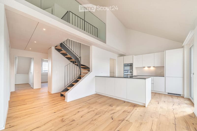 Open-plan space with wooden flooring, white walls, a modern staircase leading to an upper level, and a white kitchen area with cabinets and appliances.