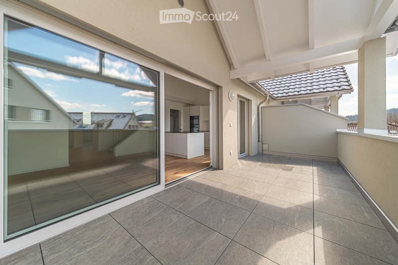 Spacious balcony with tiled flooring, large glass sliding doors leading to a modern kitchen area, and a view of neighboring houses.