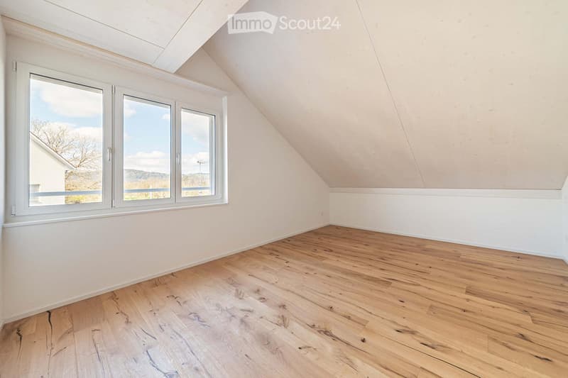Empty attic room with wooden flooring, white walls, and large windows offering a view of the outdoors.