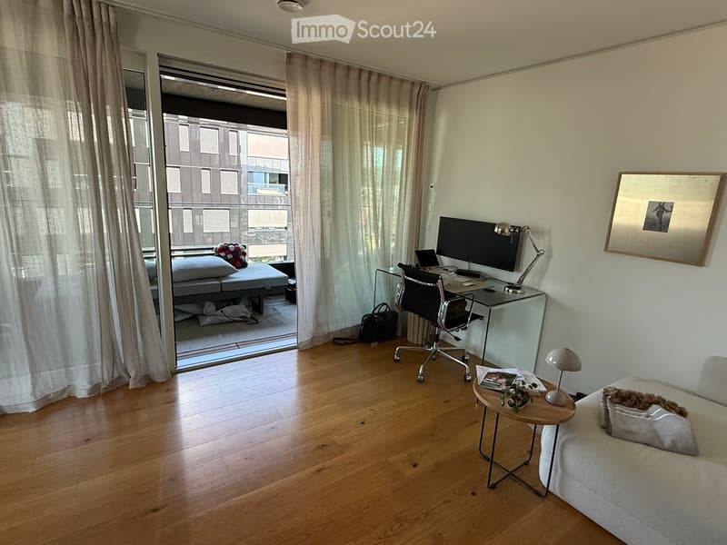 Modern furnished room with wooden flooring, large window with balcony access, sofa, desk with chair, small table with lamp, and artwork on the wall.
