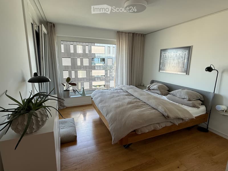 Modern bedroom with wooden flooring, large window with city view, bed with pillows and blanket, nightstand with lamp, plant, and picture frame on wall.