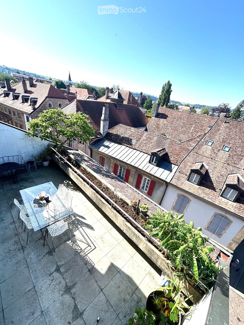 Balcony with a glass table and white chairs, overlooking a historic town with red-roofed buildings and greenery.