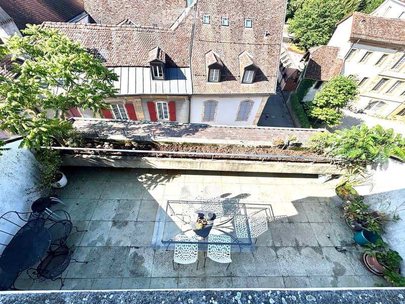 Terrace with tiled floor, glass table, chairs, and potted plants, overlooking a historic building with red shutters and tiled roof.