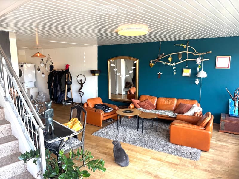 Living room with orange leather sofa, armchair, coffee table, small round table, and a cat sitting on the floor. The room features a dark blue accent wall, a large mirror, and decorative items including a hanging branch with ornaments.