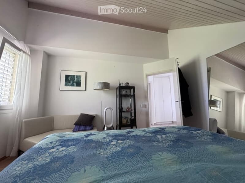 Cozy bedroom with a large bed, sofa, floor lamp, and a shelf with decorative items. The room features white walls, a window with blinds, and a mirror on the wall.