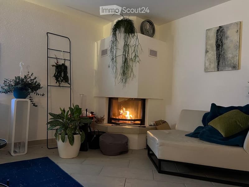 Modern living room with a white fireplace, a grey sofa with blue and green cushions, a round ottoman, a potted plant, and a decorative art piece on the wall.