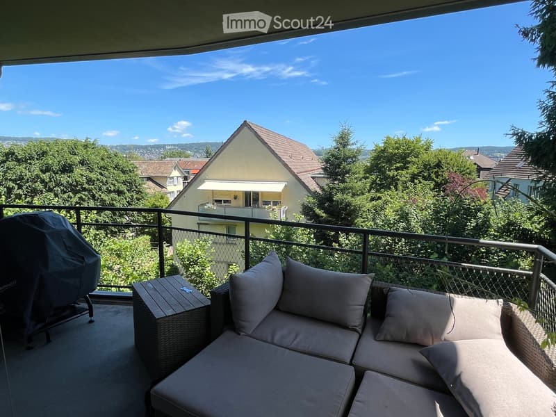 Balcony with a view of a residential area, featuring a wicker sofa with cushions and a small wooden table.