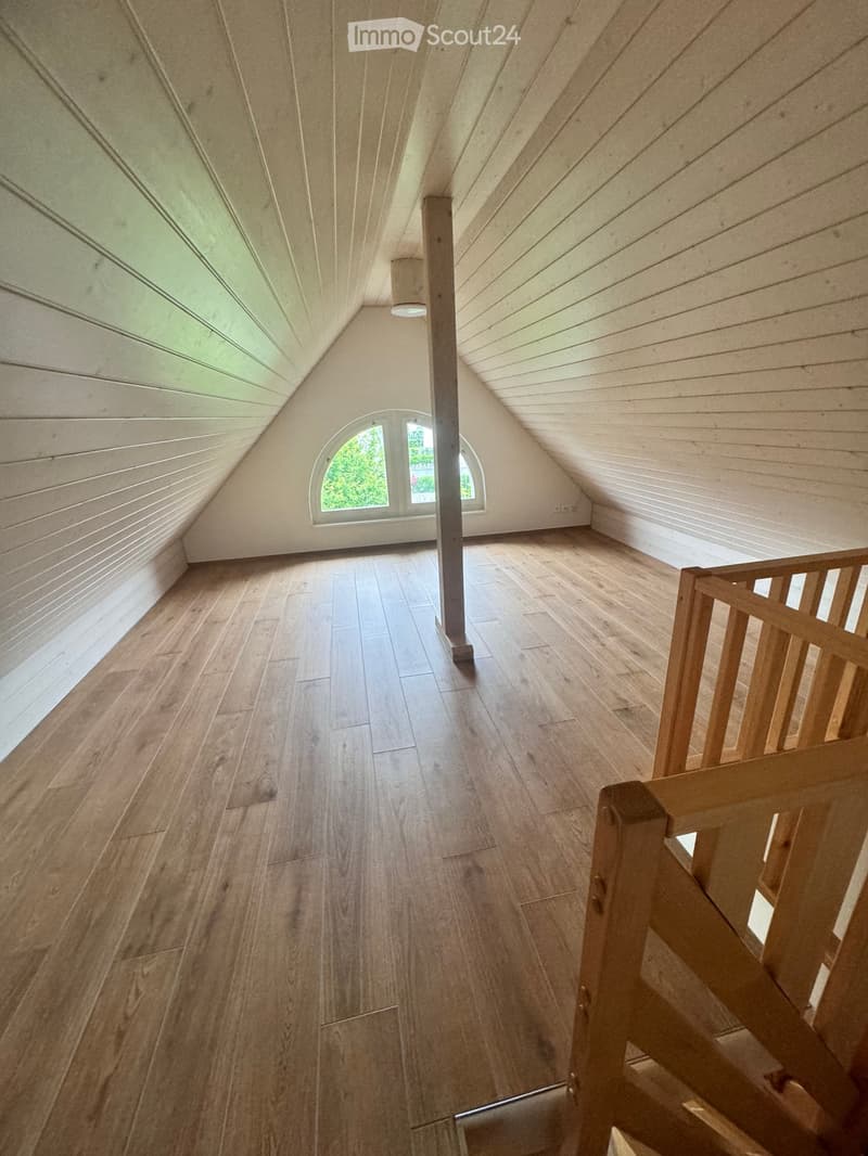 Empty attic space with wooden flooring, slanted wooden ceiling, and a small window letting in natural light.