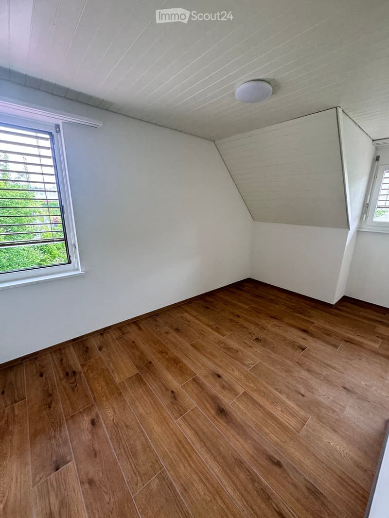 Empty room with wooden flooring, white walls, and a slanted ceiling. Features a large window with metal bars and a small window on the adjacent wall.