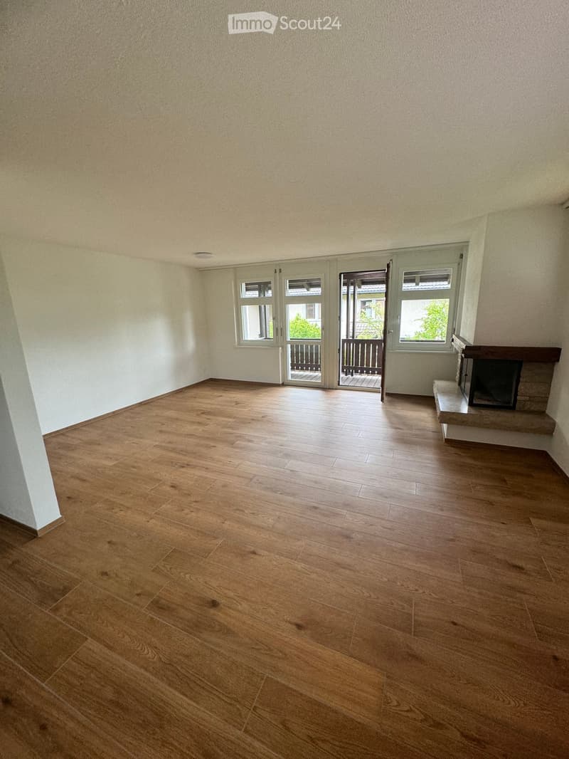 Empty room with wooden flooring, white walls, and a fireplace on the right side. There are large windows and a door leading to a balcony.