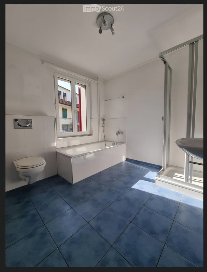 Modern bathroom with blue tiled floor, white walls, bathtub, toilet, and sink. Features a large window with a view of the street.