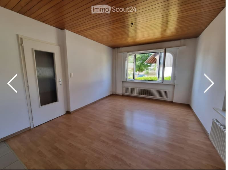 Empty room with wooden flooring, white walls, and large windows with open blinds. The room features a wooden ceiling and a radiator under the windows.