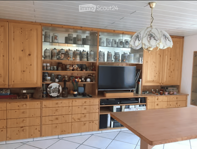 A well-furnished room with wooden cabinets, a glass display cabinet filled with jars and decorative items, a wooden table, a television, and various decorative pieces including a flower vase and a decorative plate.