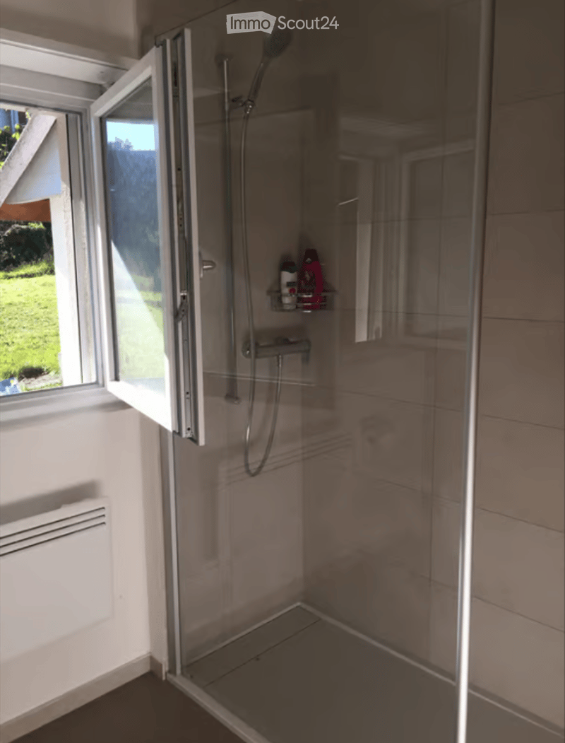Modern walk-in shower with glass enclosure, showerhead, and a small shelf holding toiletries. The shower has a large window offering a view of the garden.