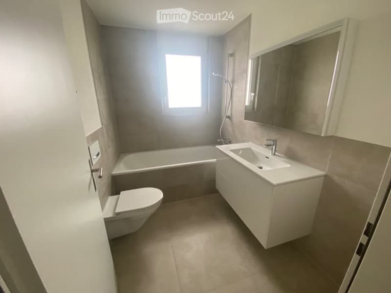 Modern bathroom with a white sink, bathtub, toilet, and a large mirror above the sink. The walls are tiled in neutral tones, and the floor is covered with beige tiles.