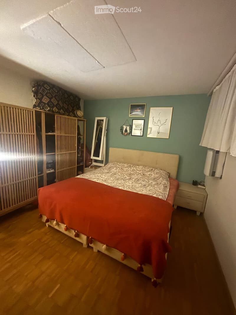 A cozy bedroom with a wooden floor, a large bed with a red blanket, a wardrobe, a mirror, and several picture frames on the wall.