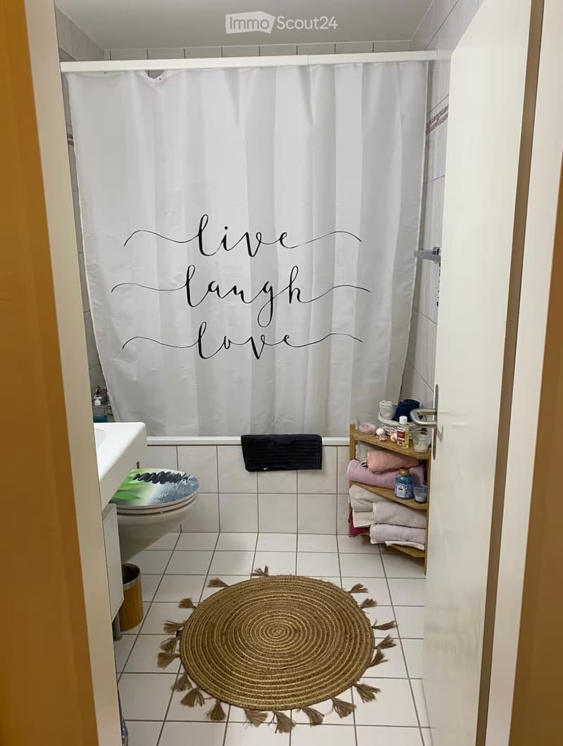 A small bathroom with white tiled walls and floor, a toilet, a sink, a shower curtain with the words "live laugh love" written on it, a towel rack with towels, and a round woven bath mat with tassels.
