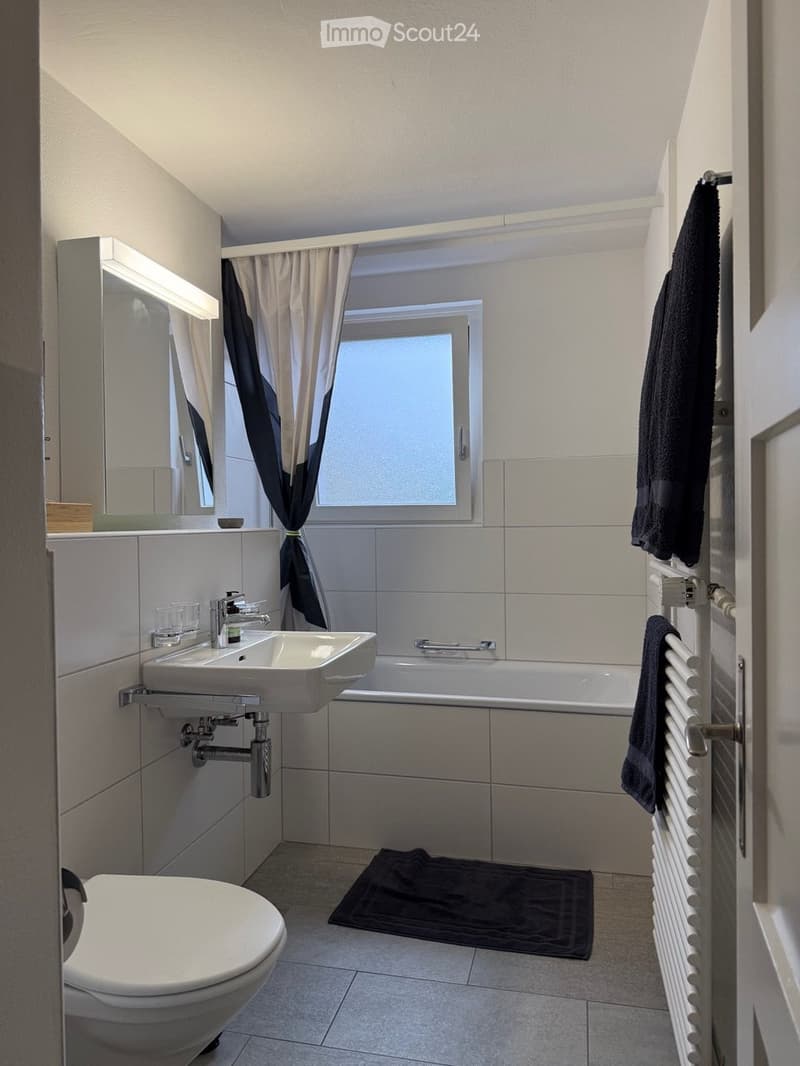 Modern bathroom with a white sink, bathtub, toilet, mirror, radiator, and a window with curtains. Towels are placed on the radiator and floor.
