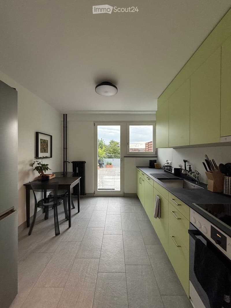 Modern kitchen with light green cabinets, stainless steel appliances, and a dining area with a table and chairs. The space features tiled flooring, a large window, and a door leading to a balcony.