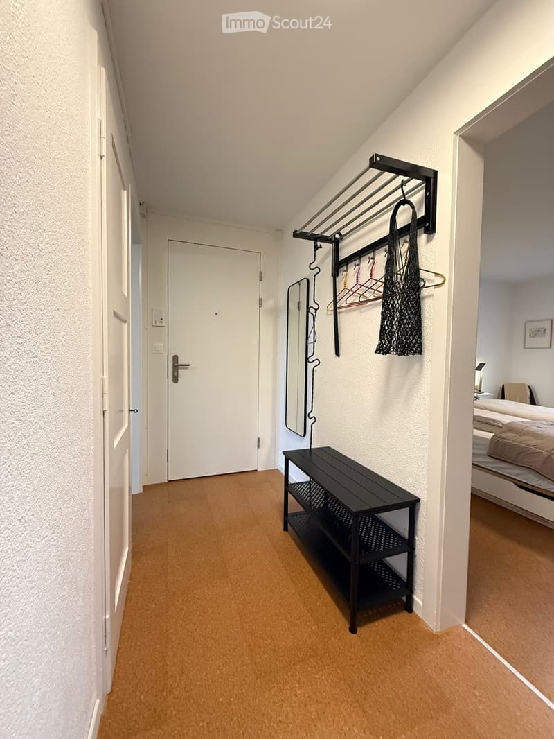 A narrow hallway with light-colored walls and carpeted floor, featuring a black bench, coat rack, and a glimpse of a bedroom with a bed and nightstand.