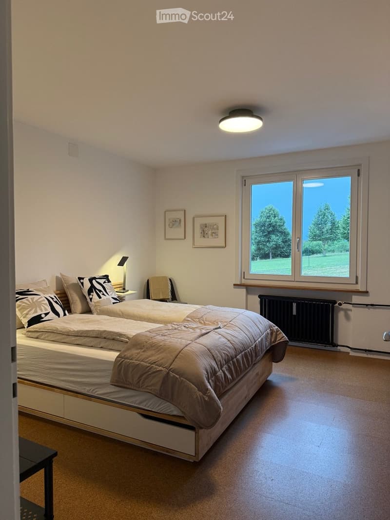 Bright bedroom with two single beds, nightstand, lamp, chair, radiator, and framed pictures on the wall. Large window offers a view of greenery.