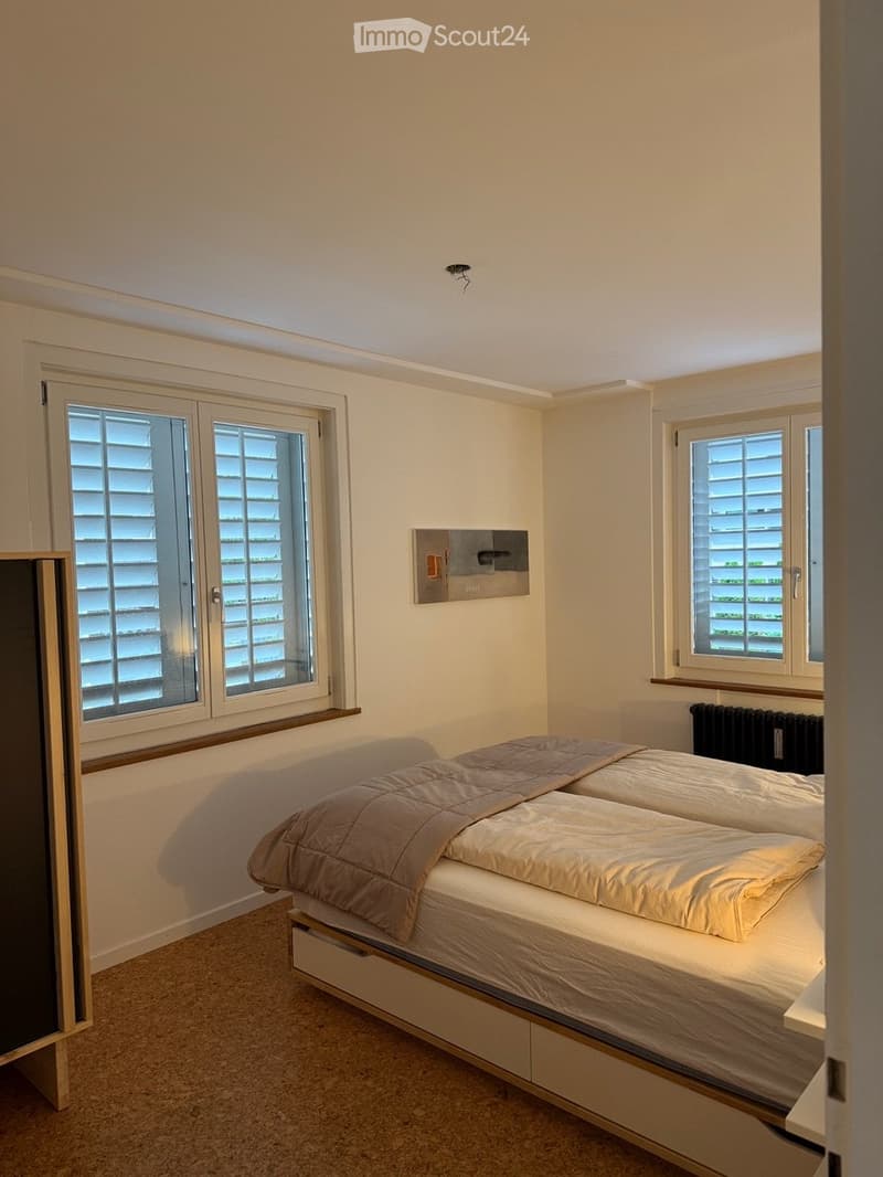 A tidy bedroom with a double bed, beige bedding, and two windows with shutters. The room has light-colored walls and carpeted flooring.