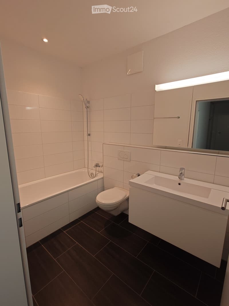 Modern bathroom with white tiles, dark tiled floor, bathtub, toilet, sink, mirror, and showerhead.