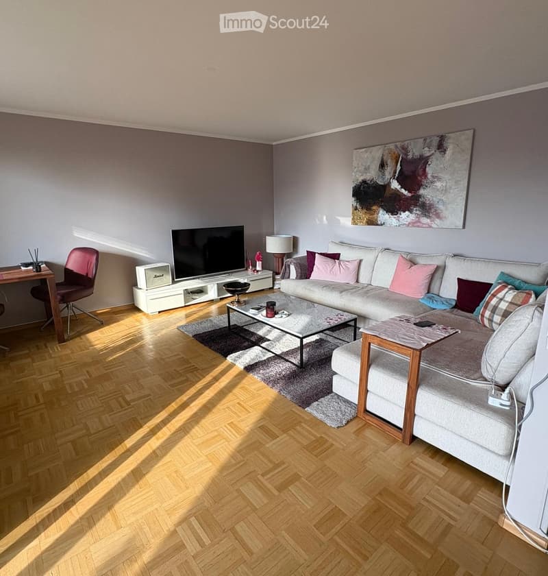 Modern living room with a grey sectional sofa, coffee table, side table, TV, and a wooden desk with a chair. The room features wooden flooring and a large abstract painting on the wall.