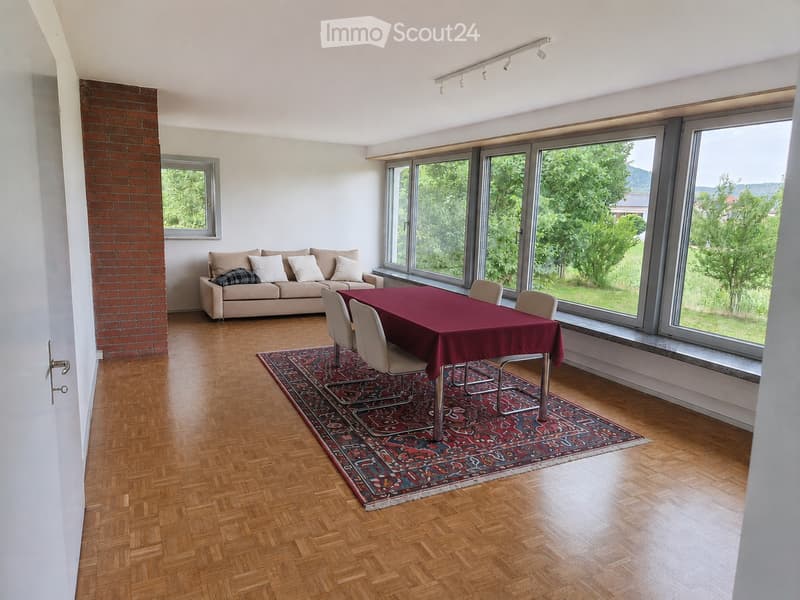 Bright living room with large windows, wooden flooring, a sofa, a dining table with chairs, and a patterned rug.