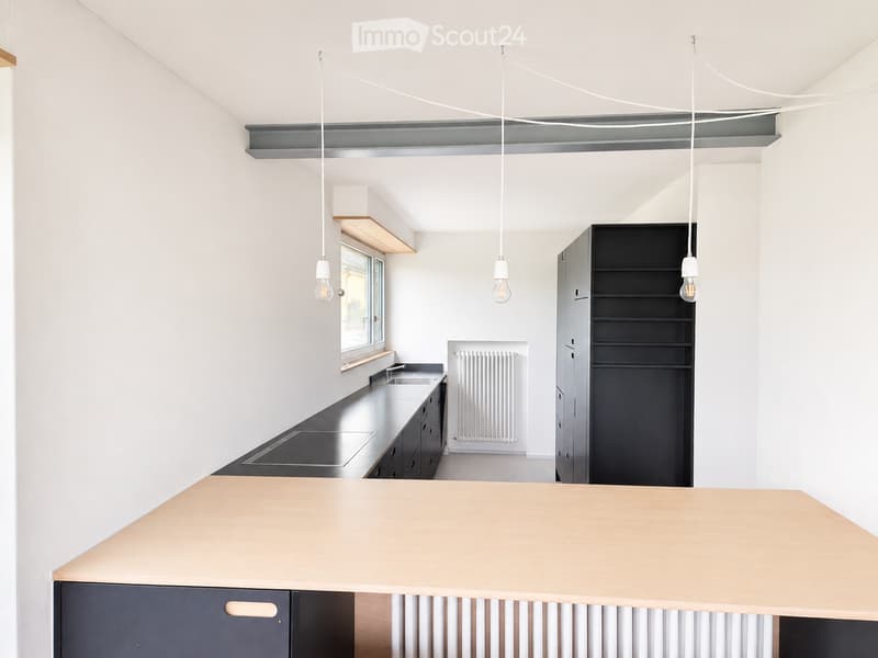 Modern kitchen with white walls, black countertops, and a light wood island. Features pendant lights, a sink, stove, and built-in cabinets.