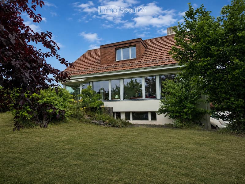 A two-story house with a red-tiled roof and large windows, surrounded by a well-maintained lawn and trees.