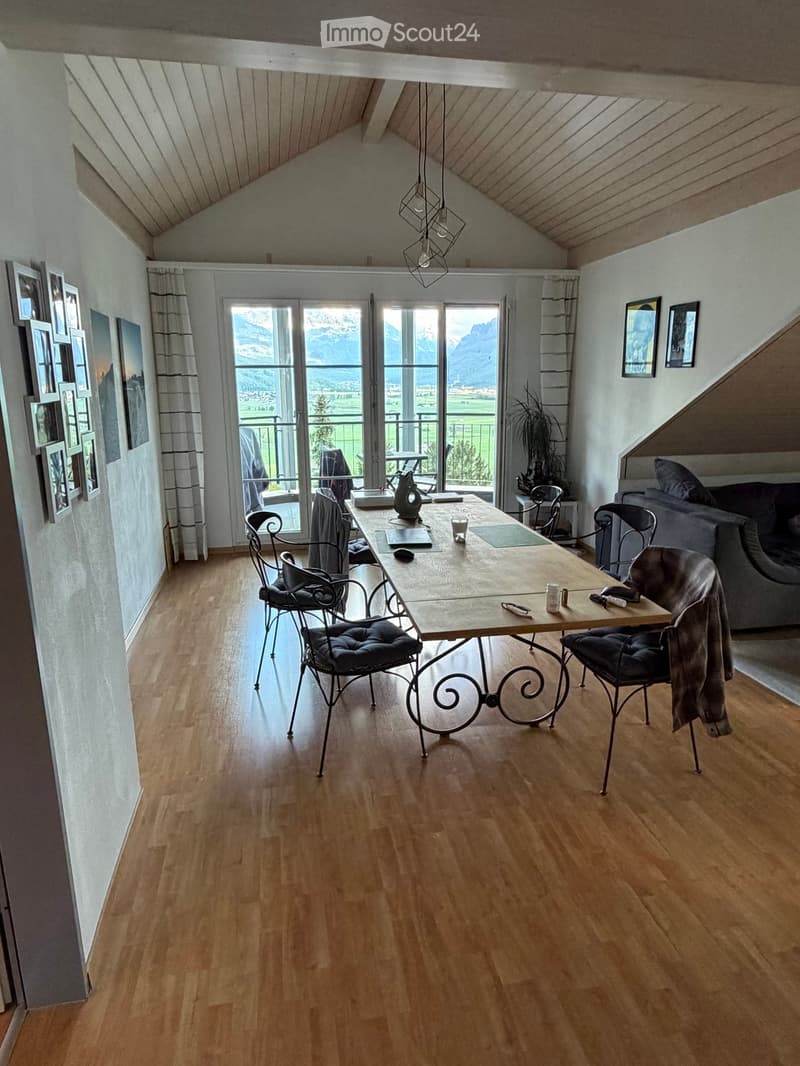 Bright living room with wooden flooring, large windows offering a scenic mountain view, a sofa, a dining table with chairs, and decorative picture frames on the walls.