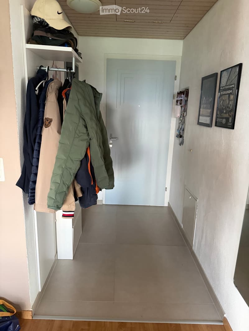A narrow hallway with a coat rack holding jackets and coats, a white door at the end, and framed pictures on the wall.