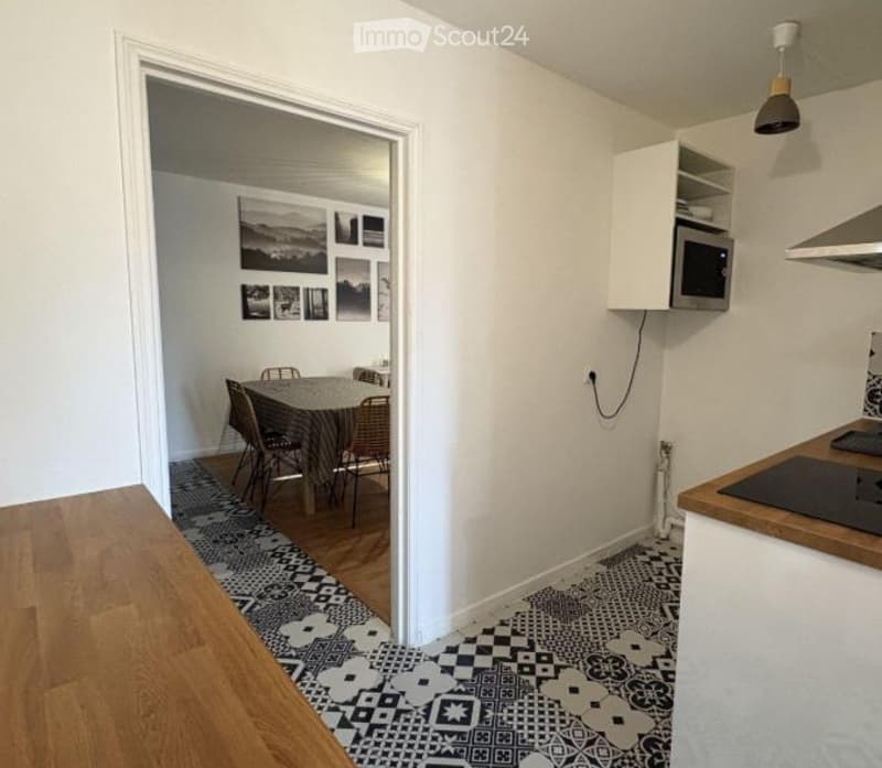 A modern, open-plan living and dining area with a dining table and chairs, a small kitchen area with a microwave, cabinet, and induction stove, and patterned tiles on the floor.