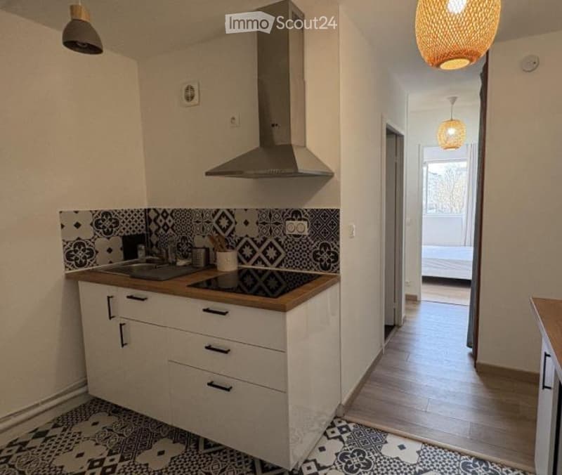 Modern kitchen with white cabinets, induction stove, and a visible bedroom area with a bed and wooden flooring.