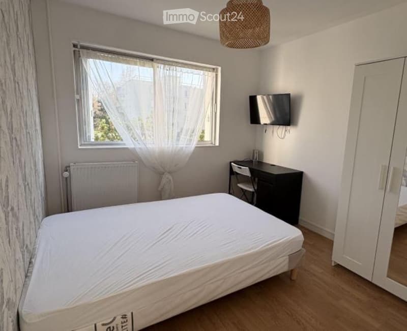 A small, modern bedroom with a single bed, a desk with a chair, a wall-mounted TV, and a large window with white curtains.
