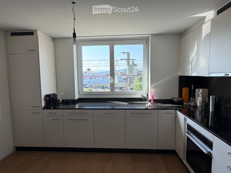 Modern kitchen with white cabinets, black countertops, and a large window overlooking the city. Includes a sink, faucet, oven, coffee maker, and various small items on the counter.